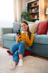 Obraz premium A cheerful Caucasian woman engages in a video call, comfortably seated on the floor, leaning against a couch in a cozy living room setting.