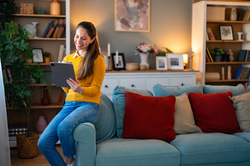A joyful woman in casual attire interacts with her digital tablet, sipping coffee as she relaxes on...