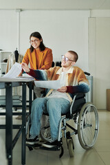Young smiling male fashion designer with disability sitting in wheelchair and discussing sketches...