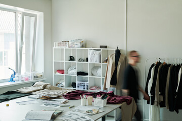 Corner of spacious modern atelier or workshop of fashion designers with variety of supplies on workplace and blurry male tailor