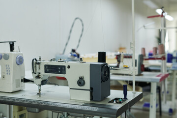 Row of desks with electric sewing machines and other tailoring equipment standing in spacious modern atelier or workshop