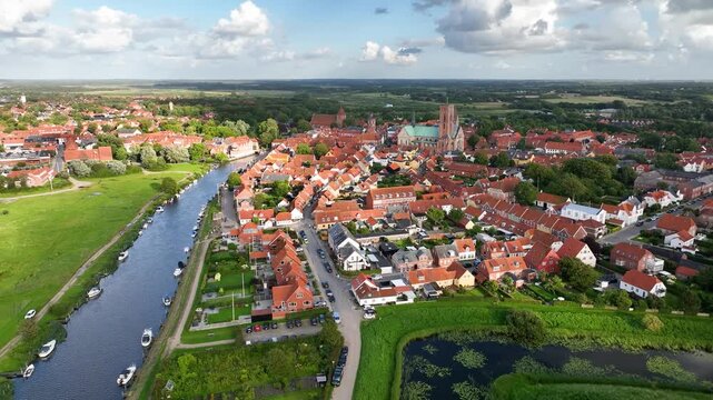 Reverse shot, moving backward to reveal the historic town of Ribe, with the majestic Ribe Cathedral, red-tiled roofs, boats docked along the riverside, and Riberhus Castle Bank, Denmark