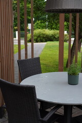 Dark rattan furniture outside after the rain. Round white table with plant pot on it. Green foliage and grass on the back. Wooden planks. Near Oru hotel. Tallinn, Estonia, Europe. July 2024