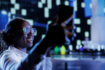 Woman making faces while using mobile phone to take selfie walking around city at night. Citizen using cellphone to take pictures of herself for social media while strolling outside on empty streets