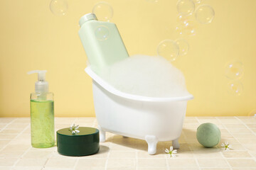 Composition with mini bathtub, bottles of cosmetic products and foam on tile table near yellow wall