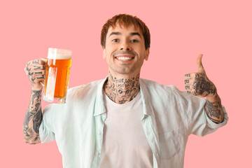 Tattooed young happy man with mug of beer showing thumb-up gesture on pink background