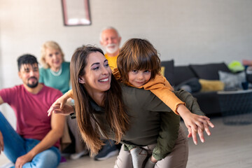 A cheerful woman enjoys piggybacking her young son, while a multi-generational, diverse family relaxes together, sitting and smiling in the cozy ambiance of a well-lit living room.