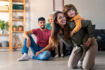 A cheerful woman piggybacks her young son while a diverse multi-generational family relaxes together on a cozy living room sofa and floor, enjoying each other's company in casual attire.