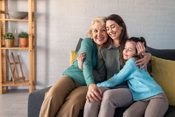 A cheerful elderly woman, her adult daughter, and young granddaughter enjoy a warm embrace, seated on a living room sofa, radiating familial love and happiness.