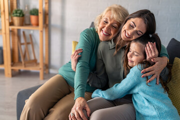 A joyful elderly woman, a smiling adult female, and a young girl enjoy a warm embrace while sitting...
