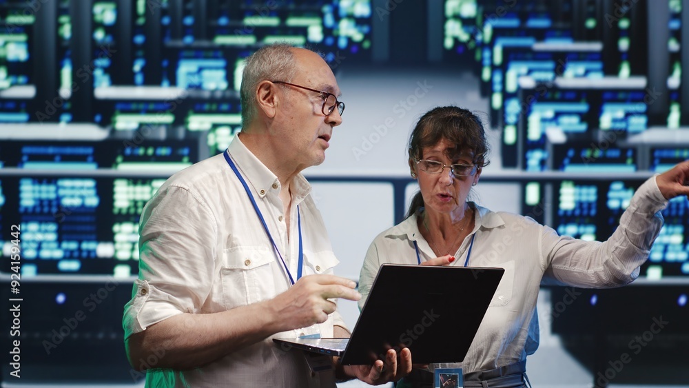 Poster seasoned tech consultants looking around server room, using laptop to crosscheck disaster recovery p