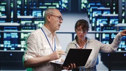 Seasoned tech consultants looking around server room, using laptop to crosscheck disaster recovery plan and assess supercomputers in need of replacement, preventing hurdles