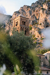 A historic stone bell tower nestled in a rugged mountainous landscape, partially obscured by lush greenery. The sun sets behind the rocky cliffs, casting a warm glow on the ancient structure.