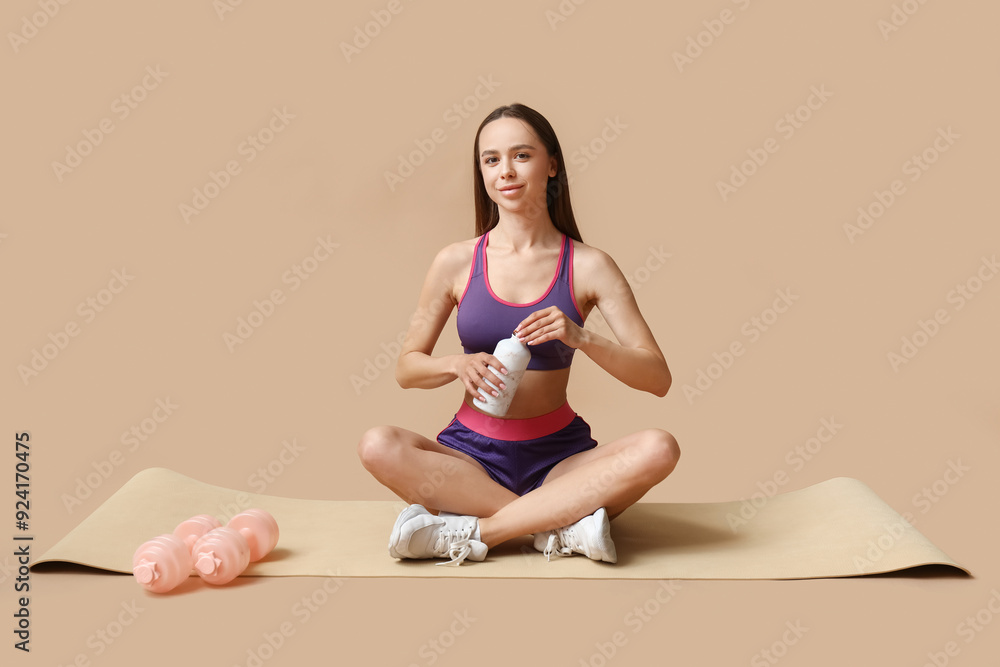 Wall mural Young female trainer drinking water and sitting on yoga mat on beige background