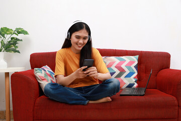 Happy young asian woman using smartphone, sitting on sofa with laptop beside her, smiling enjoying a relaxed day at home, messaging on mobile phone with friends, copy space