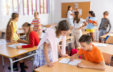 Fototapeta premium Positive friendly tween girl helping classmate sitting at school desk to complete written assignment in notebook during break before lesson ..