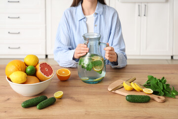 Young woman holding jug of infused water with different citrus fruits, cucumber and mint in kitchen