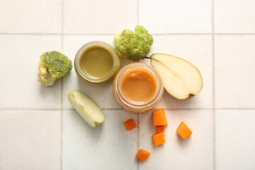 Glass jars of healthy baby food and ingredients on light tile background