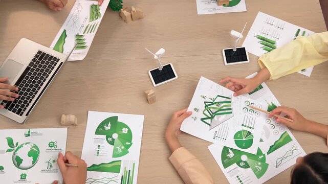 Excellent business team discussing on eco house project at meeting table with windmill design model and green power usage graph for decreasing carbon footprint, save earth. Focus on hand. Delineation.