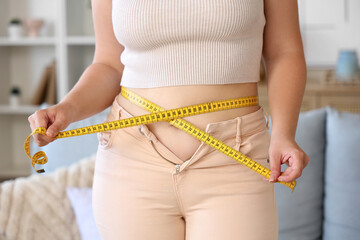 Young woman in tight pants measuring her waist at home, closeup. Weight gain concept