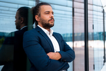Confidence successful businessman in suit with beard standing in front of office glass building lean on wall arm crossed. Hispanic handsome business man looking away. Portrait shoot side view