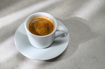 saucer and cup with milk coffee on marble gray table background with natural geometric shadow of sunlight, good morning business branding background