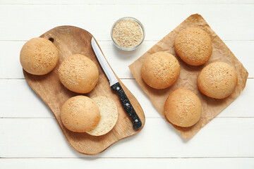 Baking paper and board of fresh buns with sesame seeds on white wooden background