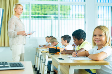 Fototapeta premium Portrait of cheerful towheaded preteen schoolgirl sitting at desk in classroom during lesson,
