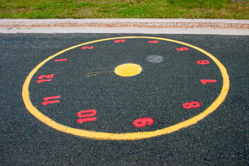 Colorful red numbers are painted on the pavement in a children's park. The school exercise game is a round yellow clock with a yellow dot in the center. The children's game helps identify time. 