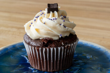 A chocolate cupcake with vanilla icing, sprinkles, and chocolate square on top. It's on a small plate and a wooden table. The homemade baked and frosted brownie-style cake's swirl is buttercream icing