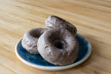 Three baked chocolate cake donuts on a round ceramic plate. There are colored sweet candy sprinkles on three homemade donuts and chocolate glaze on the fourth. The plate sits on a wooden table.