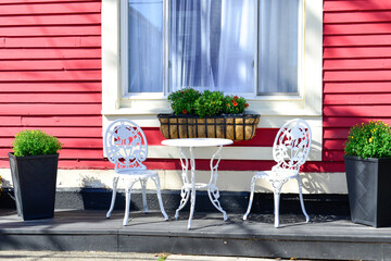 A white cast iron patio set with a round bistro table and two iron chairs. The sitting area in front of a vibrant red wooden house with clapboard siding and a large glass window has plants in pots.