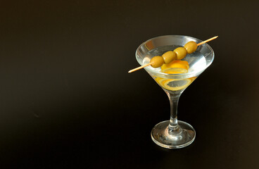 A glass of dry vermouth with ripe lemon zest and a skewer of four green olives on the edge of the vessel on a black background.