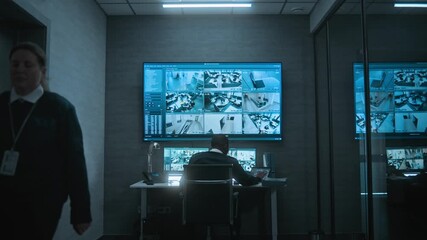 African American CCTV operator works with security cameras on computer and tablet in dim monitoring center, talks on walkie talkie. Surveillance footage on multiple monitors and big digital screen. - Powered by Adobe