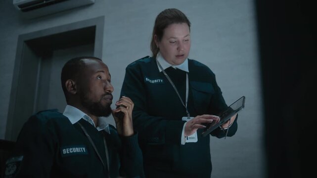 Female security officer stands and uses tablet computer. African American male dispatcher talks in walkie talkie. Multiethnic surveillance operators control CCTV cameras in modern monitoring center.