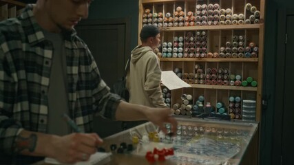 Medium close-up shot of young male graffiti shop assistant counting multicoloured plastic caps while doing inventory or stocktaking, and customer choosing spray paints for new design in background