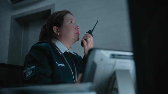 Female security officer or dispatch in uniform talks in walkie talkie, uses tablet computer and looks at screens in monitoring center during night shift. Surveillance, CCTV system and social safety.