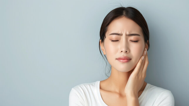 A young Asian woman with a pained expression, have a toothache and holding her cheek, with her eyes closed, standing against a light background.