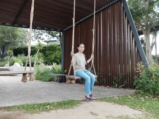 Asian woman with t-shirt and jean sitting on a swing in a park look at camera.