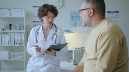 Young female doctor questioning male patient and writing notes on clipboard, both sitting on examination table during medical check-up in clinic - Powered by Adobe