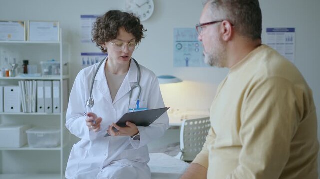 Young female doctor questioning male patient and writing notes on clipboard, both sitting on examination table during medical check-up in clinic