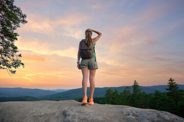 Sportive woman standing alone on hillside trail. Female hiker enjoying view of evening nature from rocky cliff on wilderness path. Active lifestyle concept