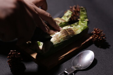 Grilled rice wrapped in banana leaf, served on brown burlap fabric with a black background, evoking...