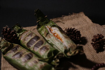 Grilled rice wrapped in banana leaf, served on brown burlap fabric with a black background, evoking...