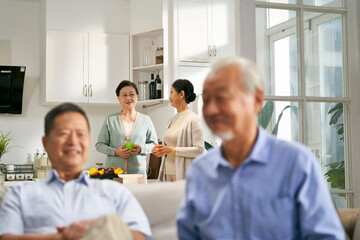two happy senior asian men sitting on couch at home having a pleasant conversation while wives chatting in kitchen, focus on background