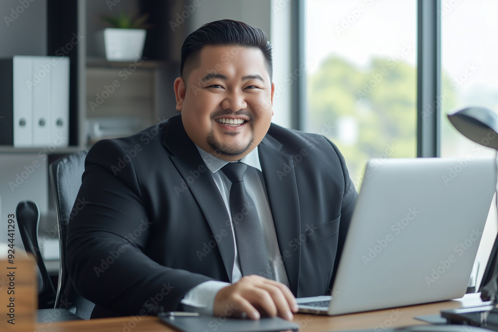 Poster asian man in a suit is sitting at a desk with a laptop in front of him. he is smiling and he is happ