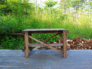 A wooden bench on the cement floor against bushes in the background. 