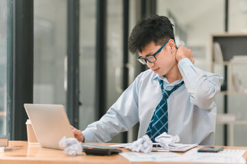 Overworked and Stressed: A young businessman sits at his desk, rubbing his neck in pain, surrounded by crumpled papers and a laptop. The image captures the exhaustion and frustration of modern office 