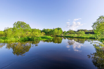 Lush trees surround a calm river, their reflections shimmering in the water under a clear blue sky, creating a tranquil atmosphere at dusk.