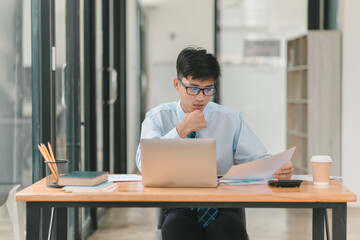 A man in a blue shirt and tie is sitting at a desk with a laptop and a piece of paper. He is looking at the paper and he is deep in thought